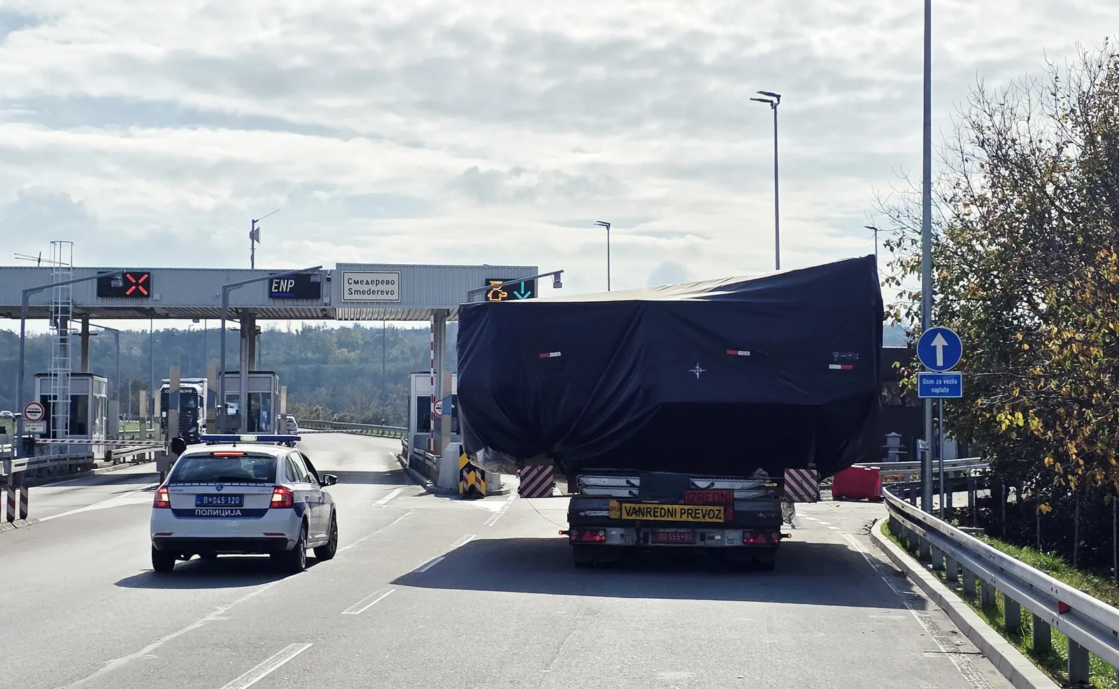 Tarpaulin-covered abnormal load passing a motorway toll plaza under police escort