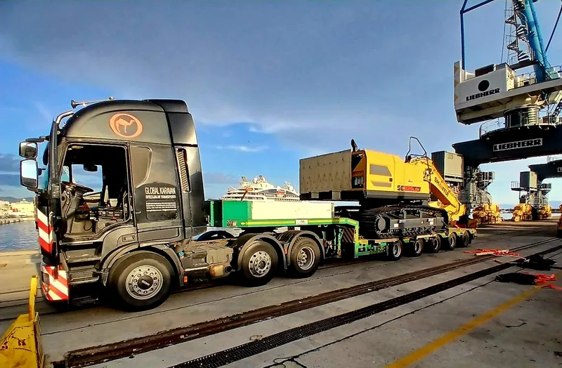 Black Iveco tractor hauling a tracked excavator on a modular semi-trailer at a port