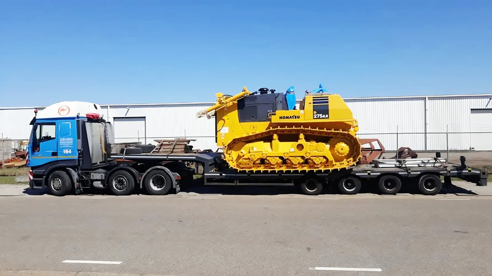 Lowboy trailer transporting a yellow Komatsu D275AX bulldozer past an industrial hall